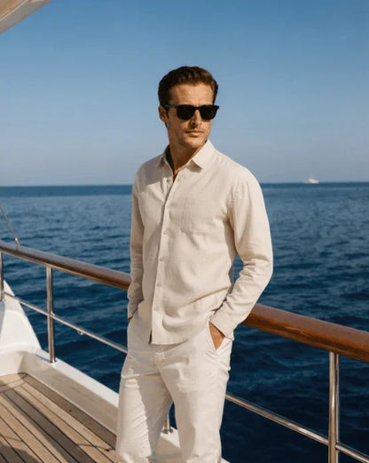 Man in white linen shirt and pants wearing sunglasses on yacht deck by the ocean