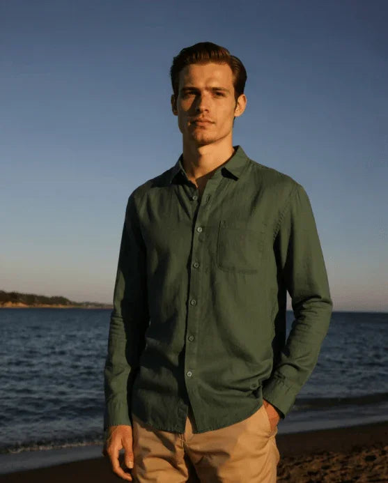 man in green button-up shirt and khaki pants standing on a beach at sunset